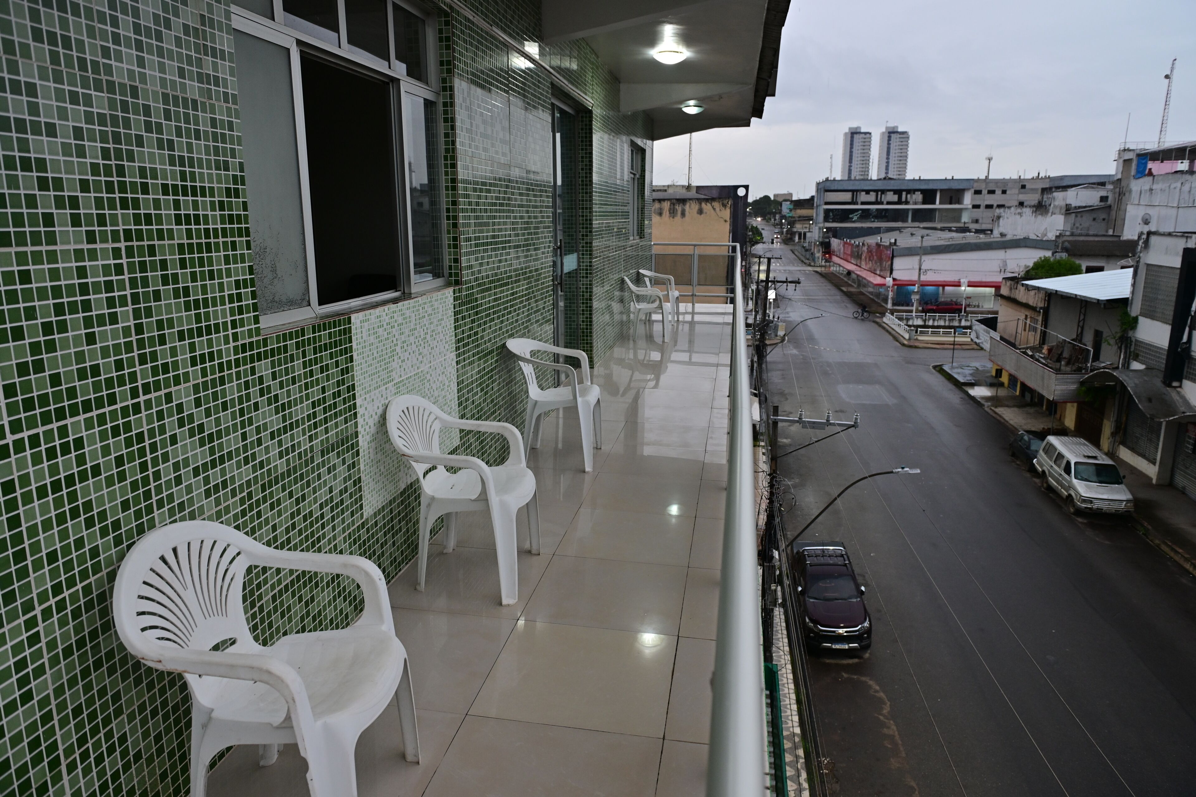 Un balcon avec des chaises en plastique, un mur recouvert de carreaux et une vue sur une rue bordée de voitures et d’immeubles.