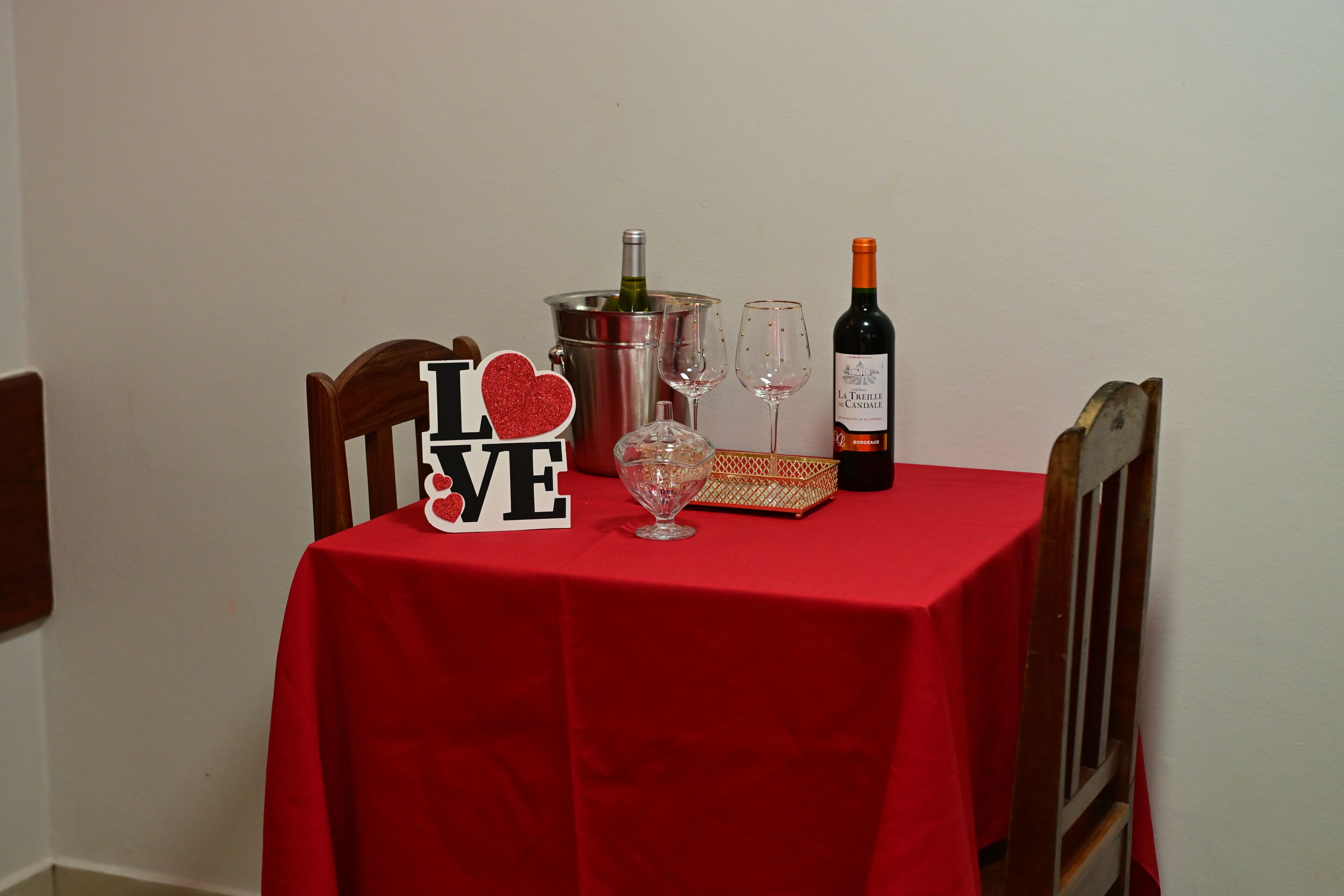 A table with a red tablecloth, a bottle of wine, two wine glasses, a champagne bottle, and a decorative sign.