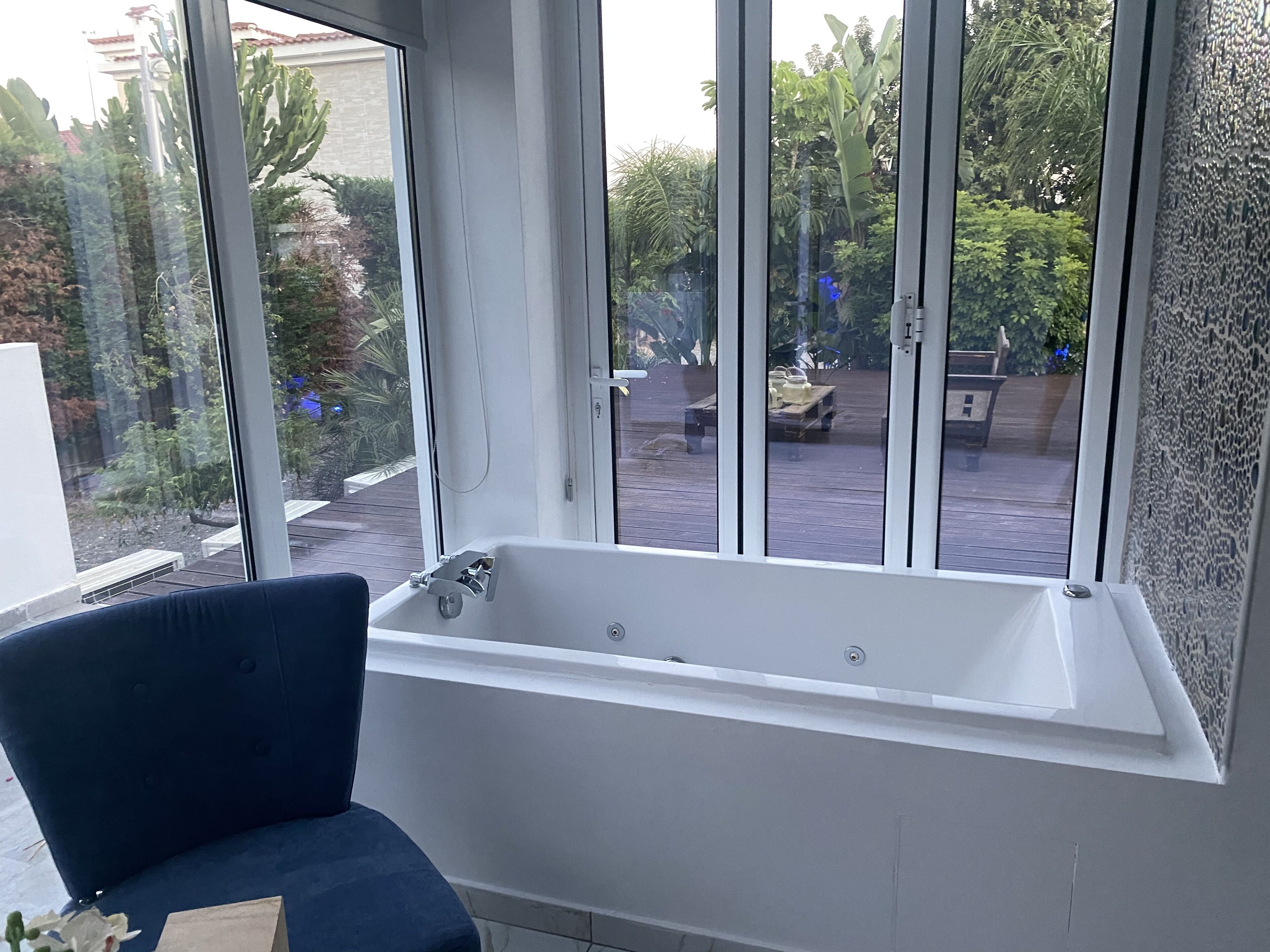 A modern bathroom with a large bathtub, a blue chair, and a view of a garden through large windows.