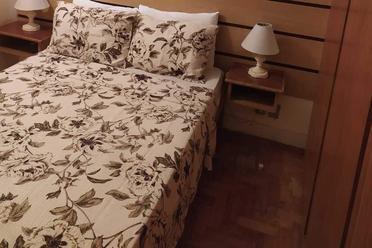 Une chambre à coucher avec une couvre-lit à motifs floraux, deux lampes de chevet, une tête de lit et un climatiseur fixé au mur.