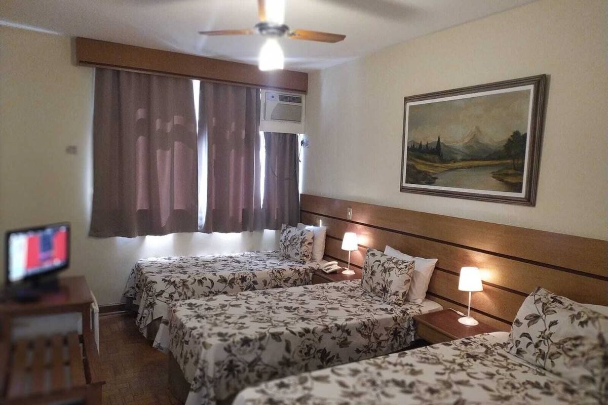 Une chambre d’hôtel avec deux lits, un ventilateur de plafond, un appareil de climatisation, un tableau au mur et un bureau avec un écran d’ordinateur.