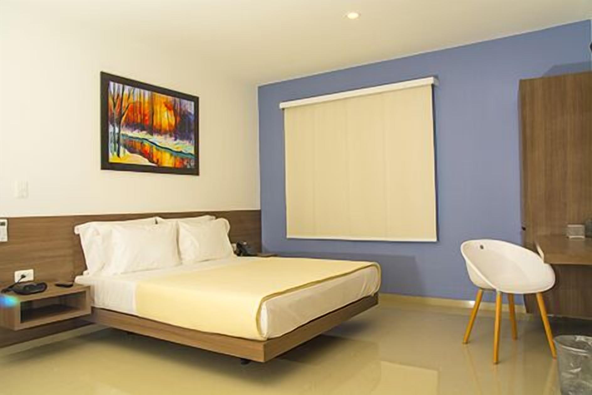 Une chambre d’hôtel avec un lit, une chaise, un tableau et une fenêtre avec des stores.
