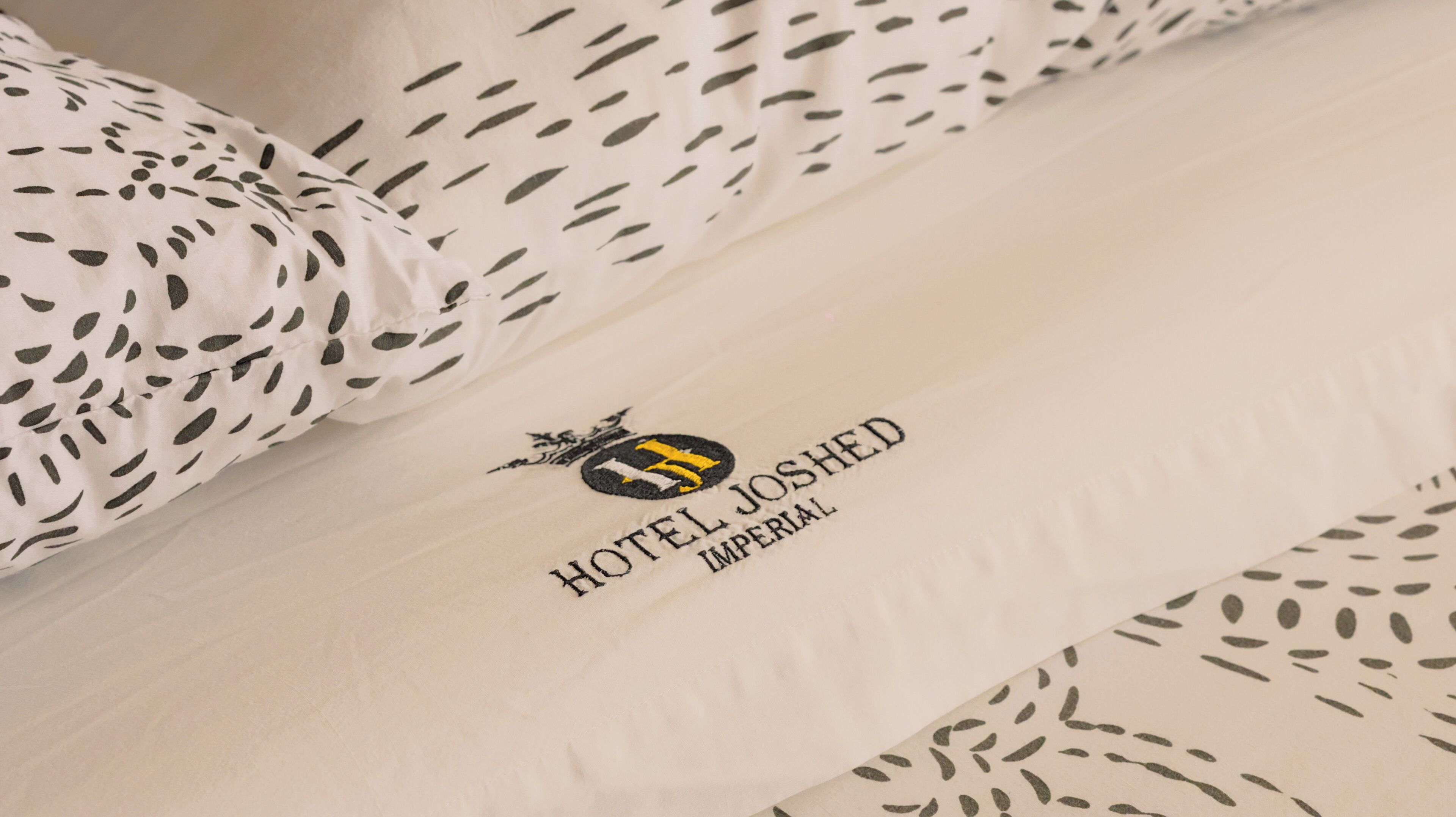 A hotel bed with a logo and name "HOTEL JOSHED IMPERIAL" on the pillowcase.