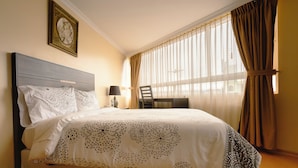 Un lit bien fait, recouvert d’une couvre-lit à motifs, une table de chevet avec une lampe, et une chaise placée près d’une grande fenêtre ornée de rideaux légers.