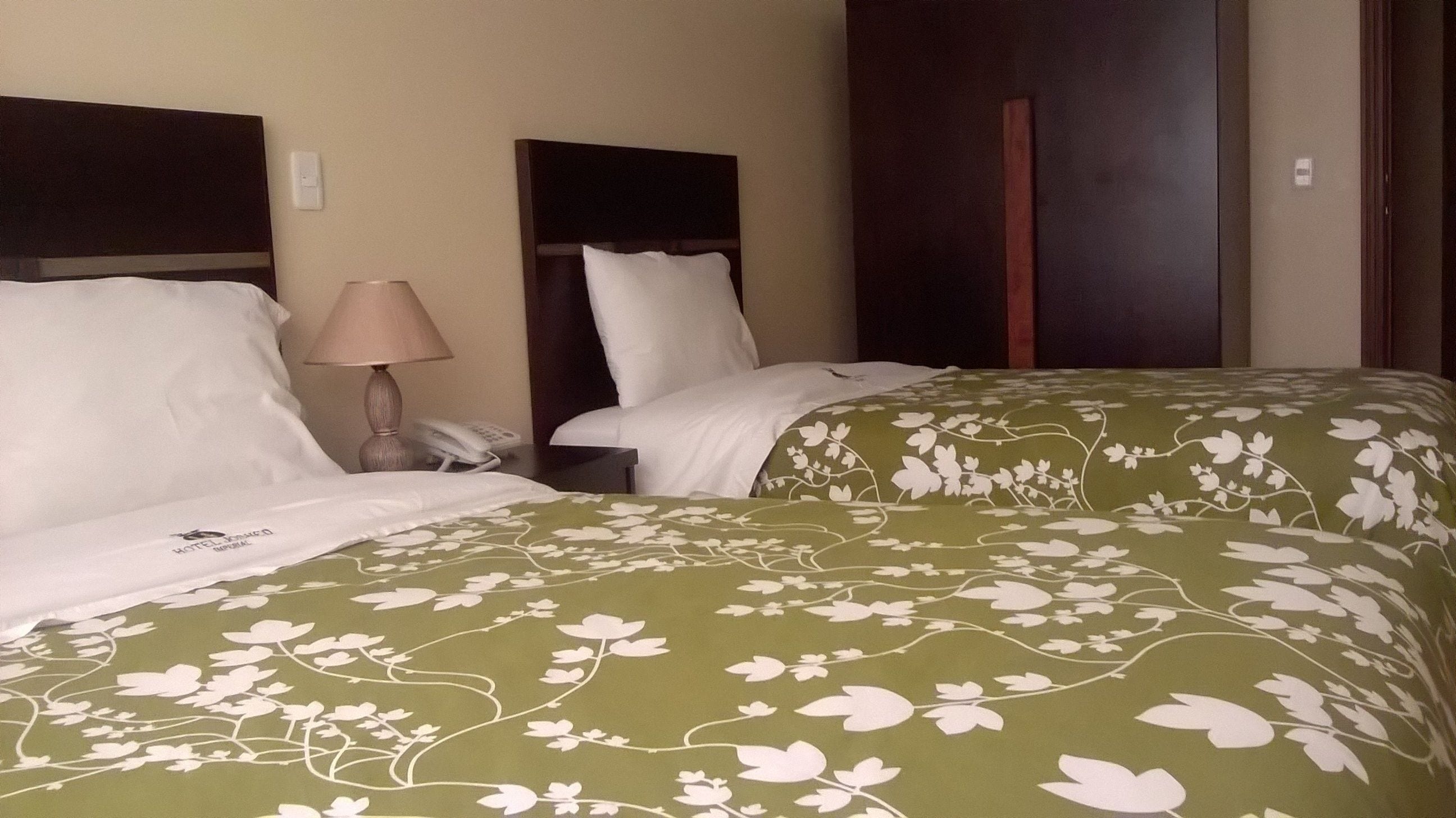 A hotel room with two beds, each with white bedding and a green floral bedspread. There is a bedside table with a lamp and a phone.
