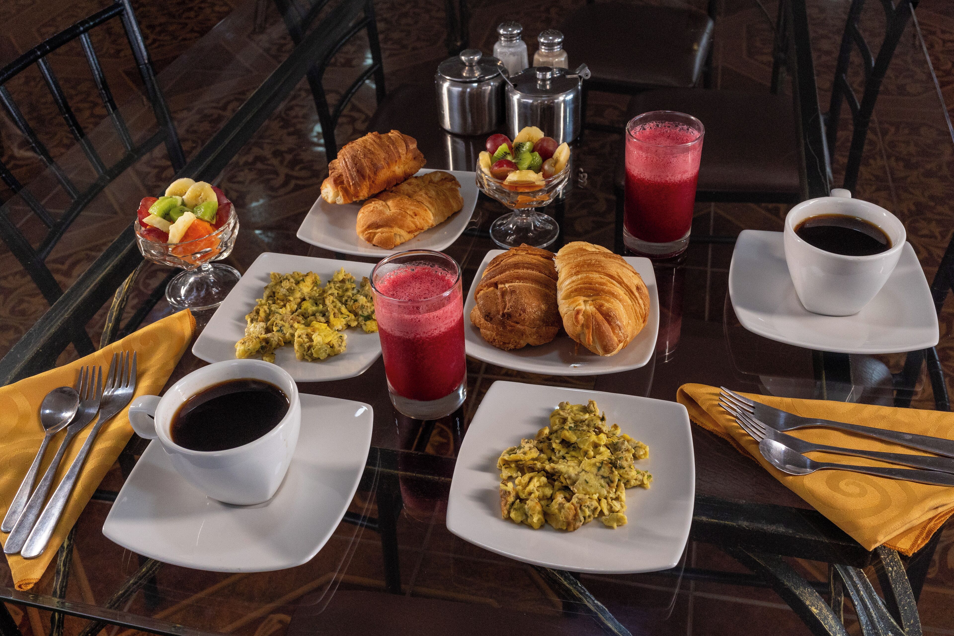 Una mesa de desayuno con cruasanes, fruta, batidos y café.