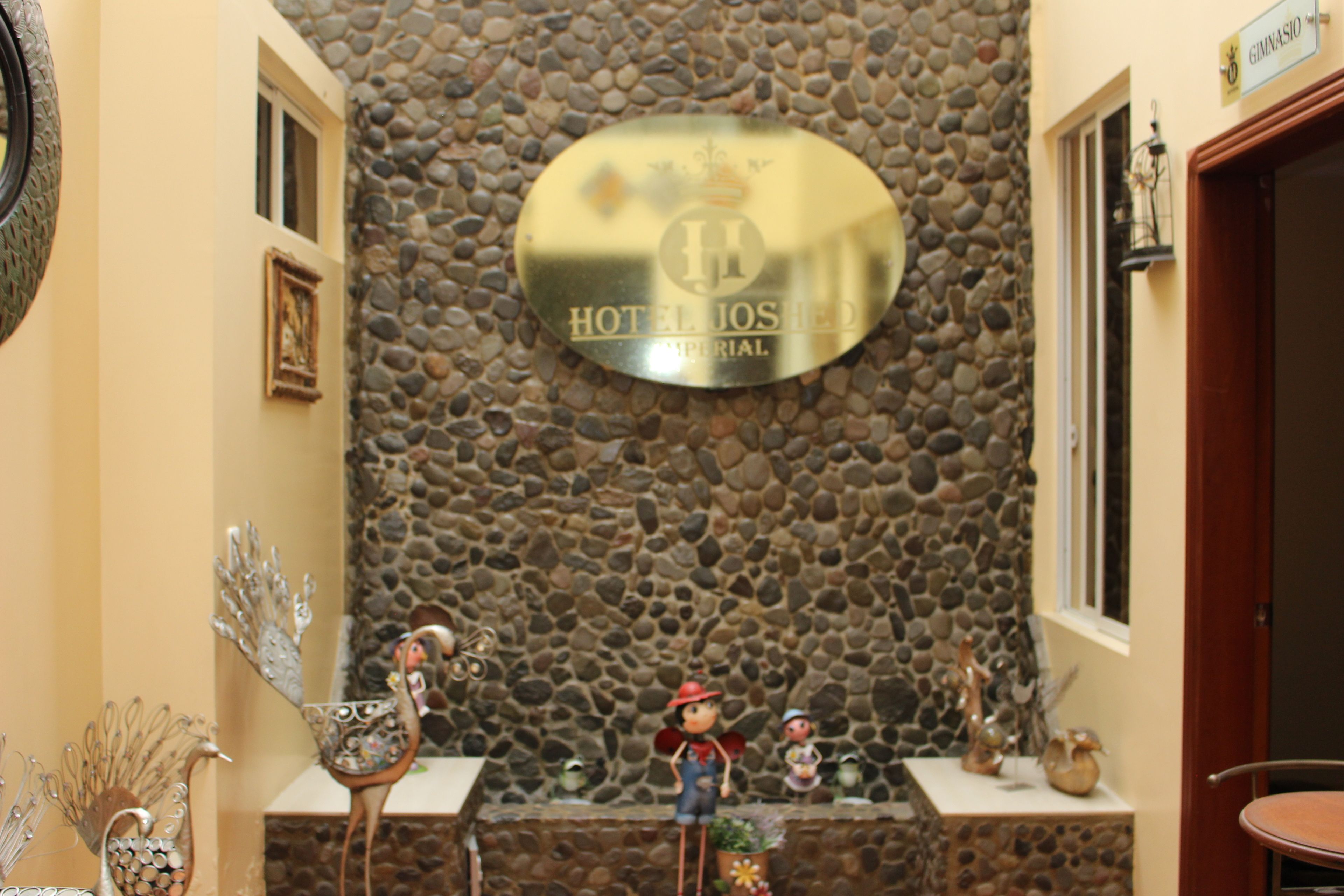Ein Empfangsbereich mit Steinwand, einem Schild mit der Aufschrift 'HOTEL JOSHEP' und Dekorationselementen, darunter ein Pfau und Figuren.