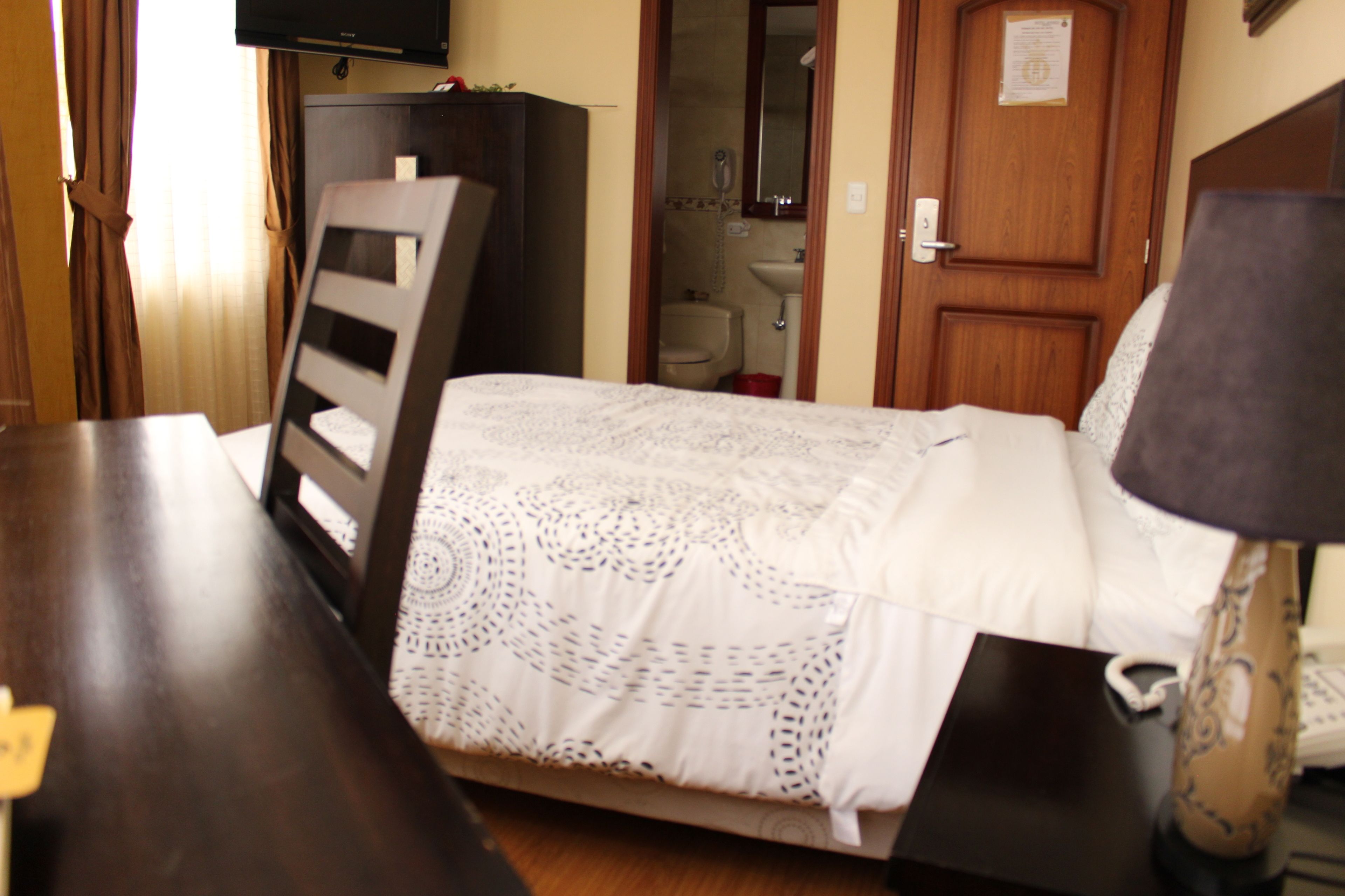 A hotel room with a bed, a wooden nightstand, a desk, a lamp, a television, and a bathroom visible through an open door.