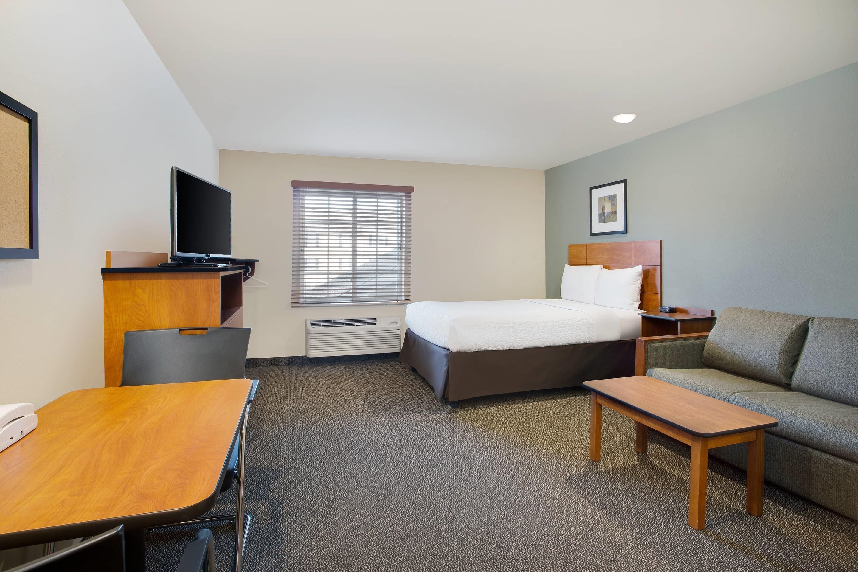 A hotel room with a bed, a desk, a television, a sofa, and a window with blinds.