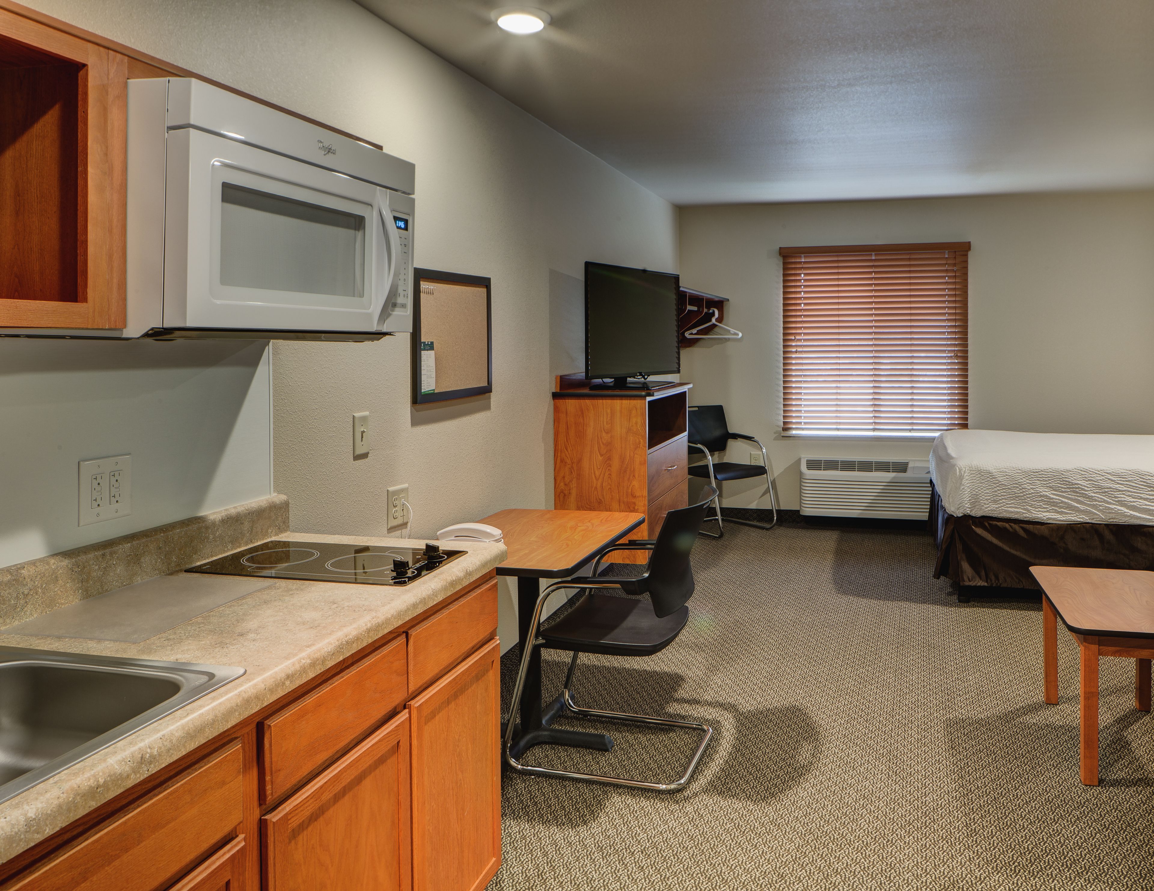 A compact hotel room with a kitchenette, a single bed, a TV, and a window with blinds.