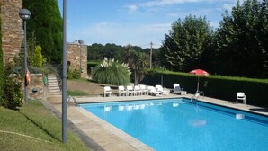 Una piscina con sillas de descanso y una sombrilla roja, rodeada de vegetación y un edificio de piedra.