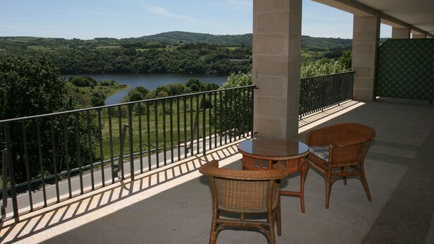 Un balcón con sillas de mimbre y una mesa, con vistas a un lago y colinas verdes.
