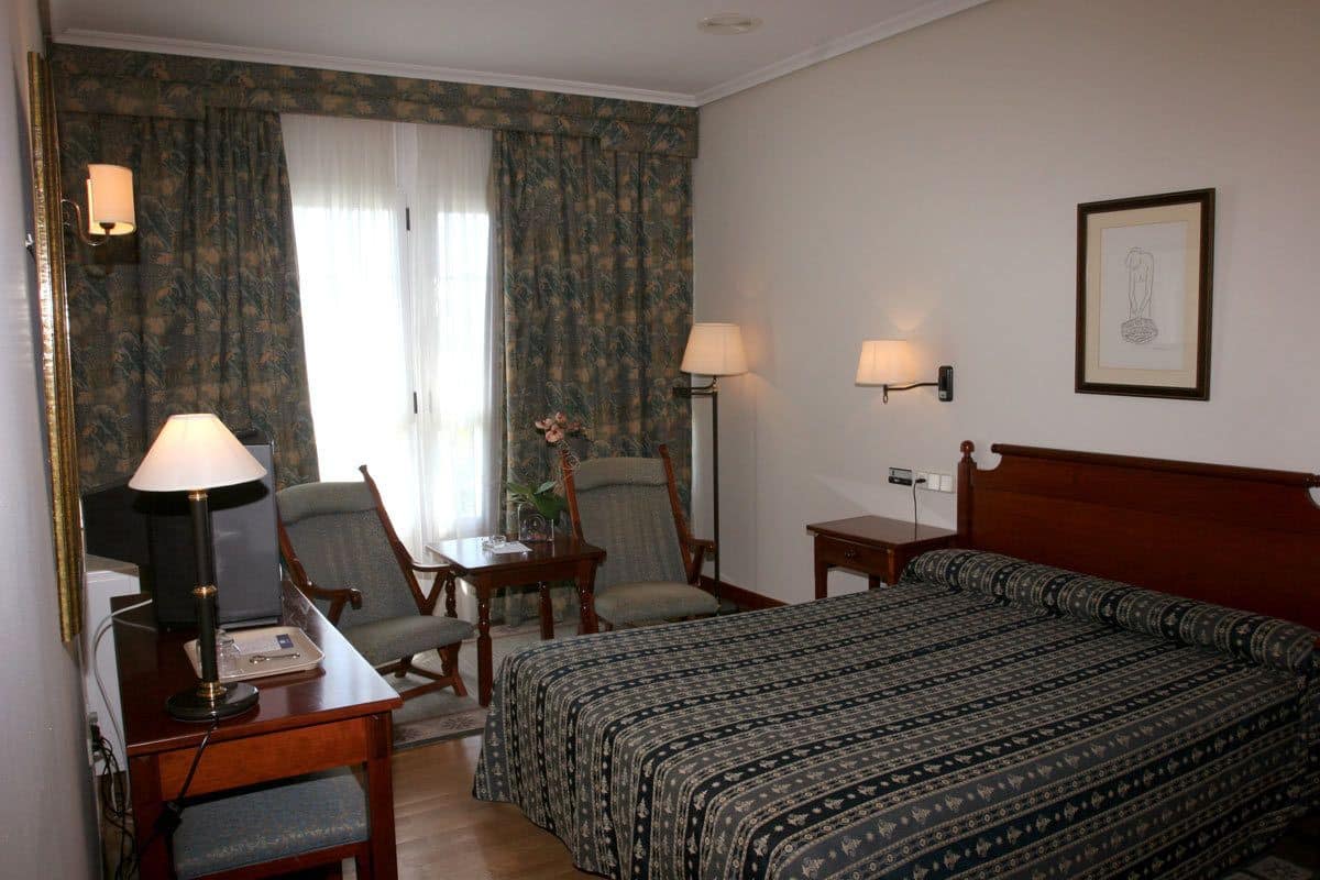 Habitación de hotel con cama, escritorio con lámpara, dos sillas, una mesita y una lámpara de pared.
