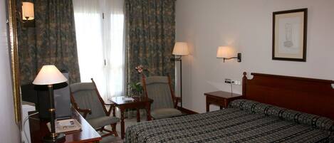 Habitación de hotel con cama, escritorio con lámpara, dos sillas, una mesita y una lámpara de pared.