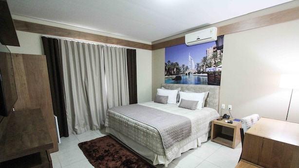 Una habitación de hotel con cama, mesita de noche, escritorio, televisor y un amplio ventanal con cortinas.