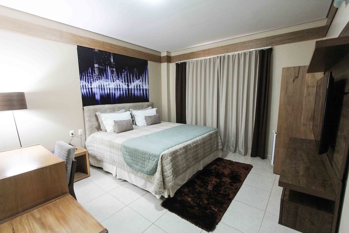 Un dormitorio con cama, escritorio, silla, televisor y una ventana grande con cortinas.