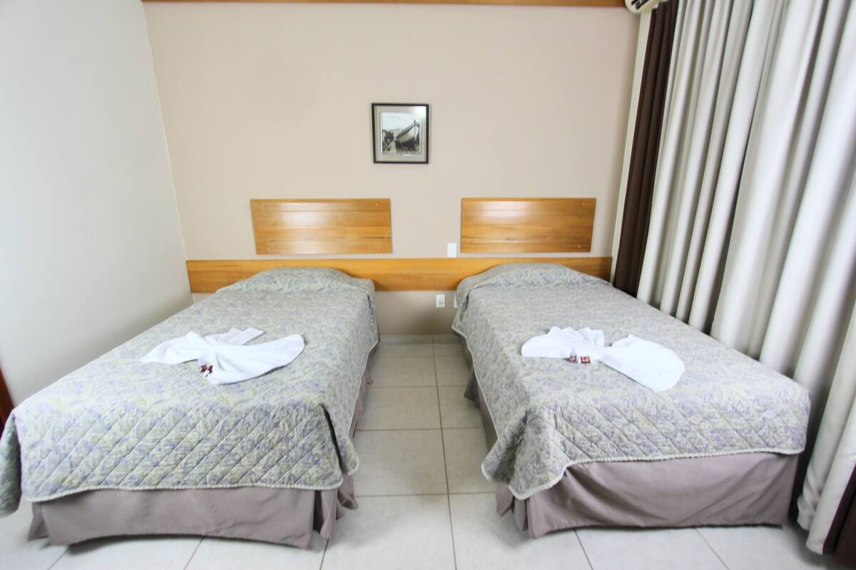 Una habitación de hotel con dos camas individuales, un cabecero de madera, un cuadro enmarcado en la pared y una ventana con cortinas.