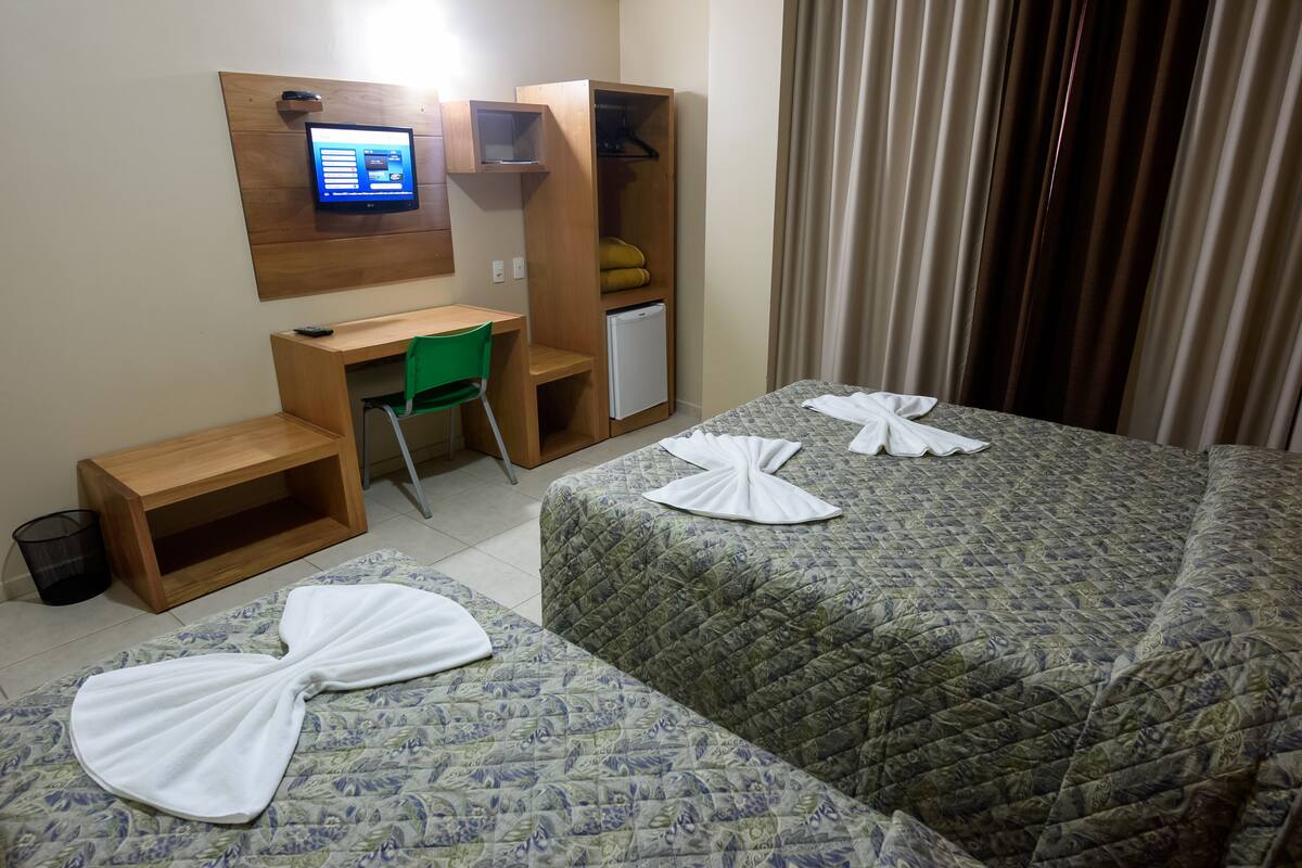 Habitación de hotel con dos camas, un escritorio con ordenador, un refrigerador pequeño y una silla verde.