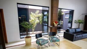 Una sala de estar moderna con un ventanal que da a un balcón, un sofá de cuero negro, dos sillones estampados, una mesa de centro de madera con un jarrón de flores rojas y una planta grande en maceta.