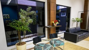 Un lobby moderno con un separador de vidrio, una recepción, un sofá y dos sillones con una planta en maceta y un jarrón de flores.
