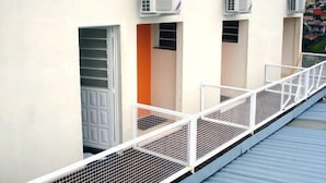 A building with three air conditioning units mounted on the exterior wall, a white metal railing along a balcony, and a view of a residential area in the background. - Pousada Estrela do Vale (Taubate)