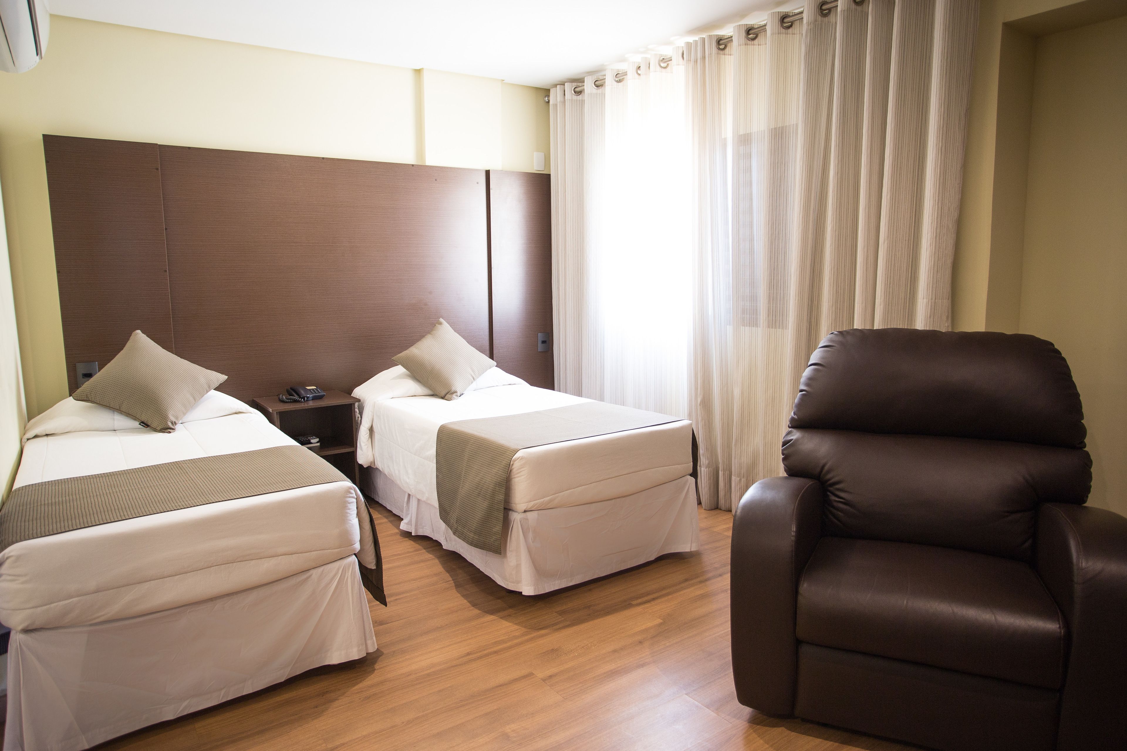 A hotel room with two beds, a brown leather armchair, and a large window with sheer curtains.