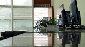 Un mostrador de recepción con superficie de vidrio, dos monitores, una planta en maceta y un cartel que indica las tarifas de las habitaciones.
