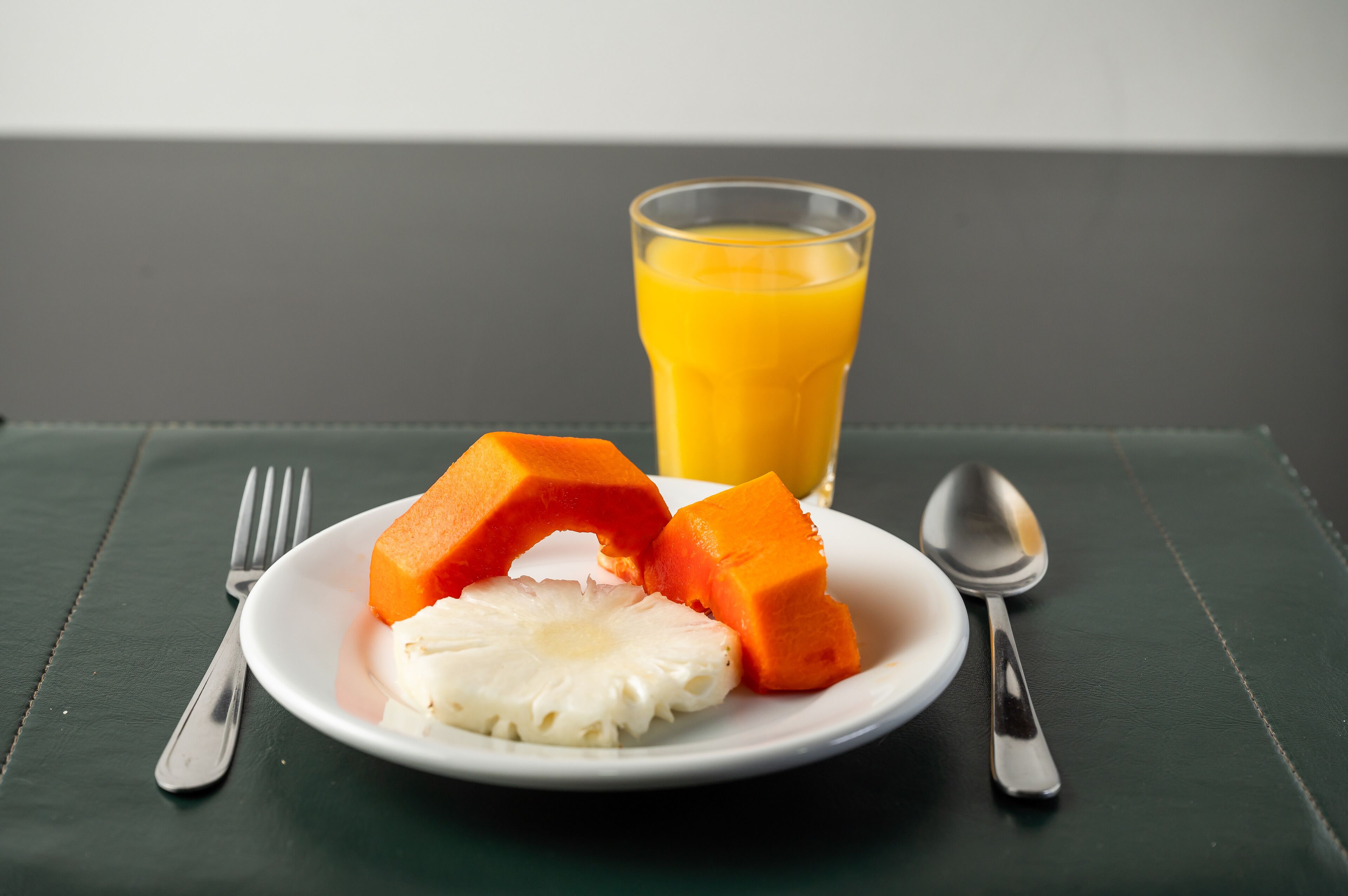 Un plato con piña y papaya sobre un mantel oscuro, acompañado de un vaso de jugo de naranja y cubiertos.