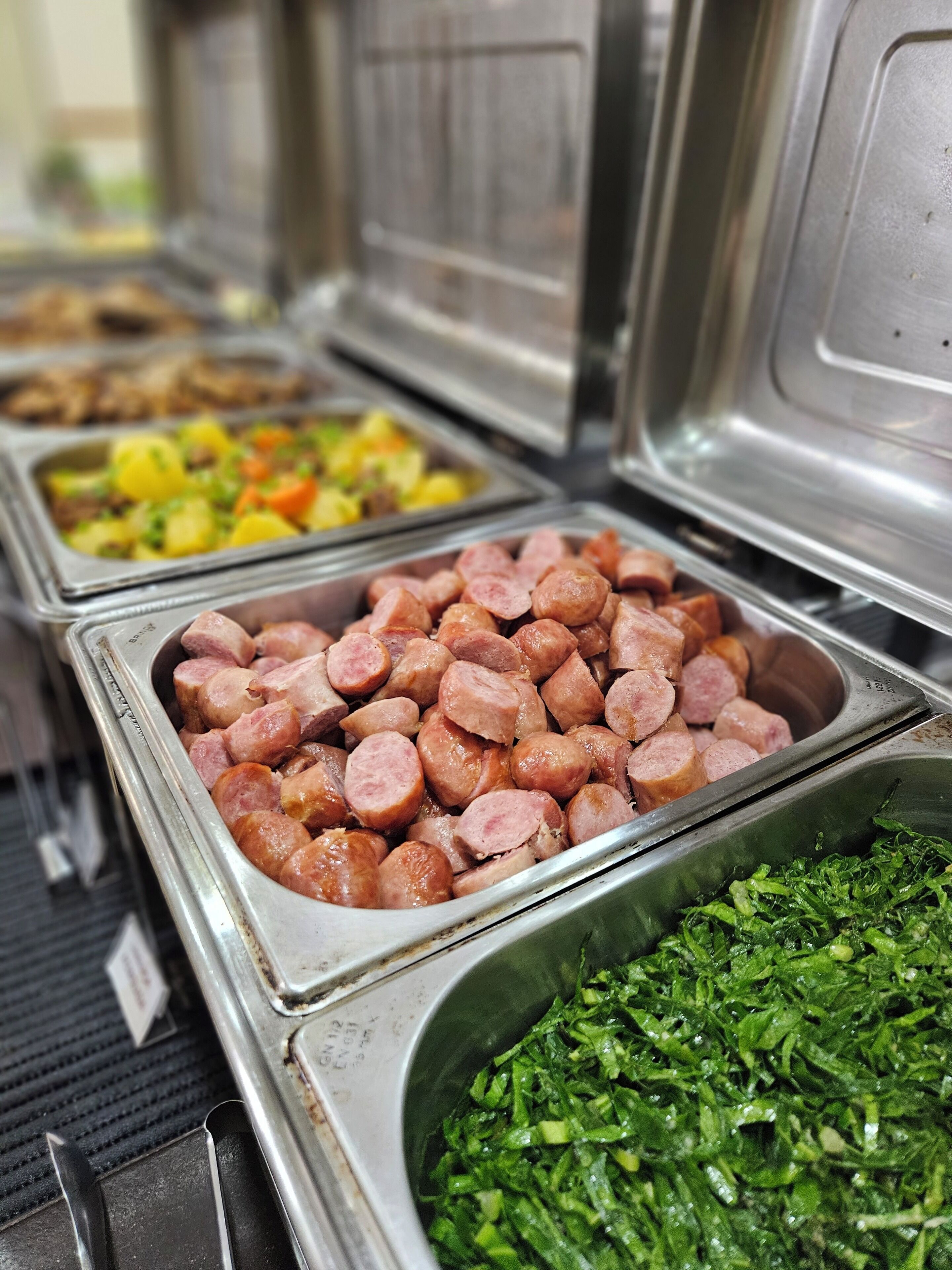 Un bufé con diversas opciones de comida, incluyendo salchichas en rodajas, un plato de papas y verduras, y un recipiente con hojas verdes picadas.