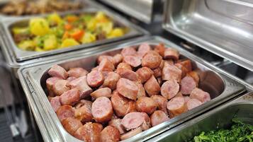 Un bufé con diversas opciones de comida, incluyendo salchichas en rodajas, un plato de papas y verduras, y un recipiente con hojas verdes picadas.