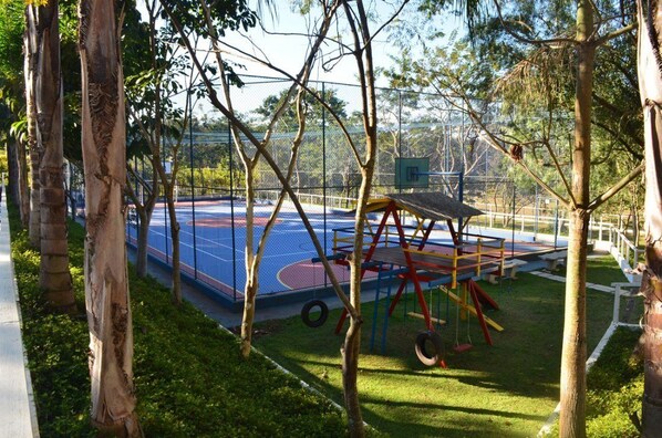 Una cancha de baloncesto rodeada de árboles y un parque infantil con columpios y tobogán.