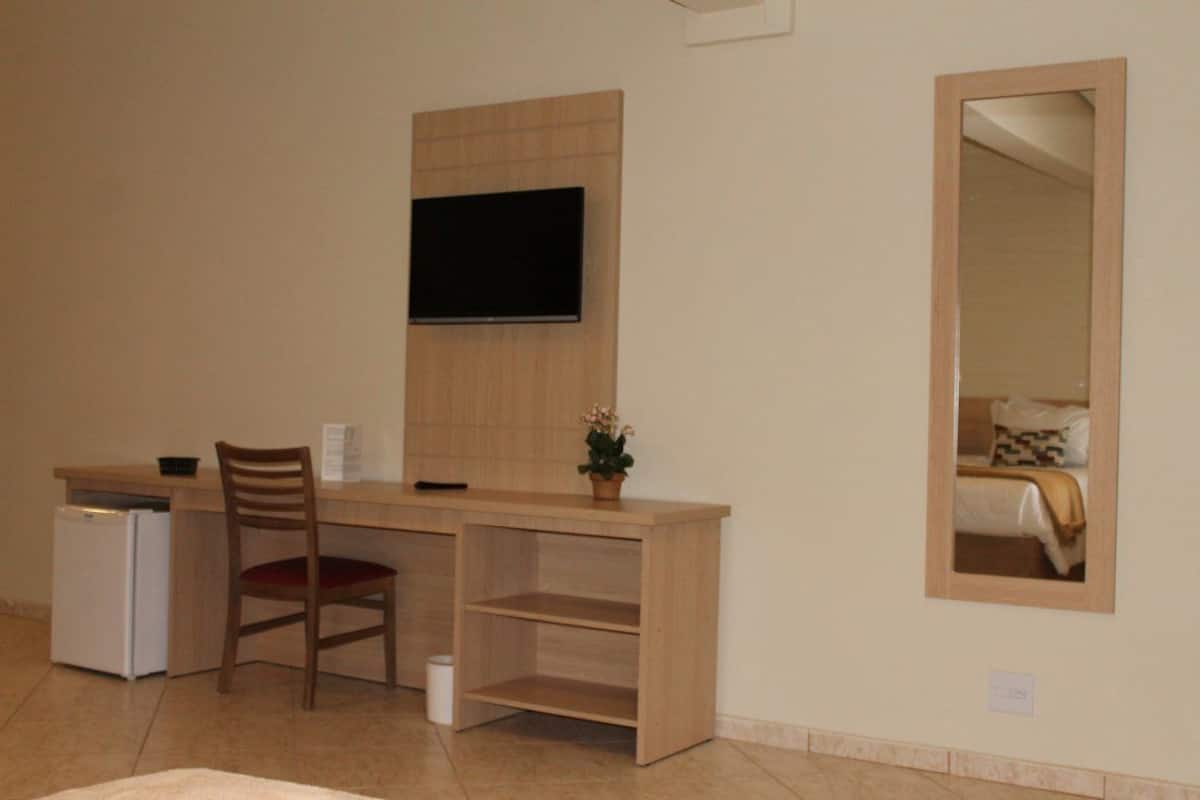 A compact room with a wooden desk, a chair, a small refrigerator, a television mounted on the wall, and a mirror reflecting a bed.