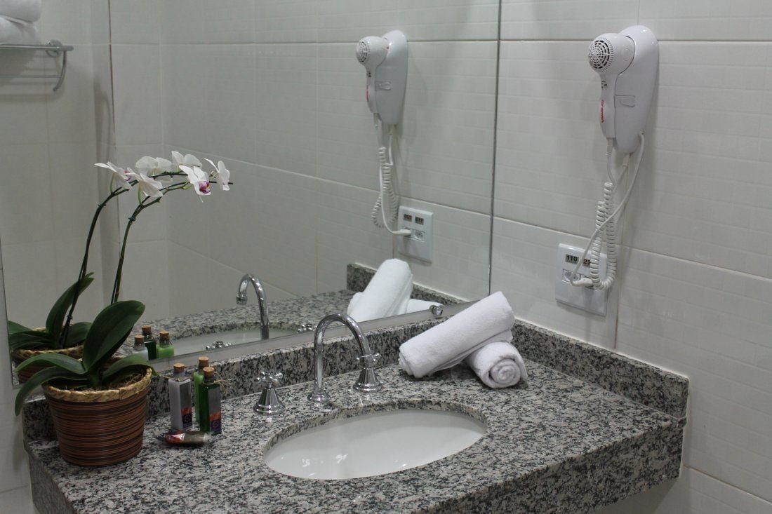 A bathroom with a granite sink, a mirror, two hair dryers mounted on the wall, a potted orchid, and toiletries on the counter.
