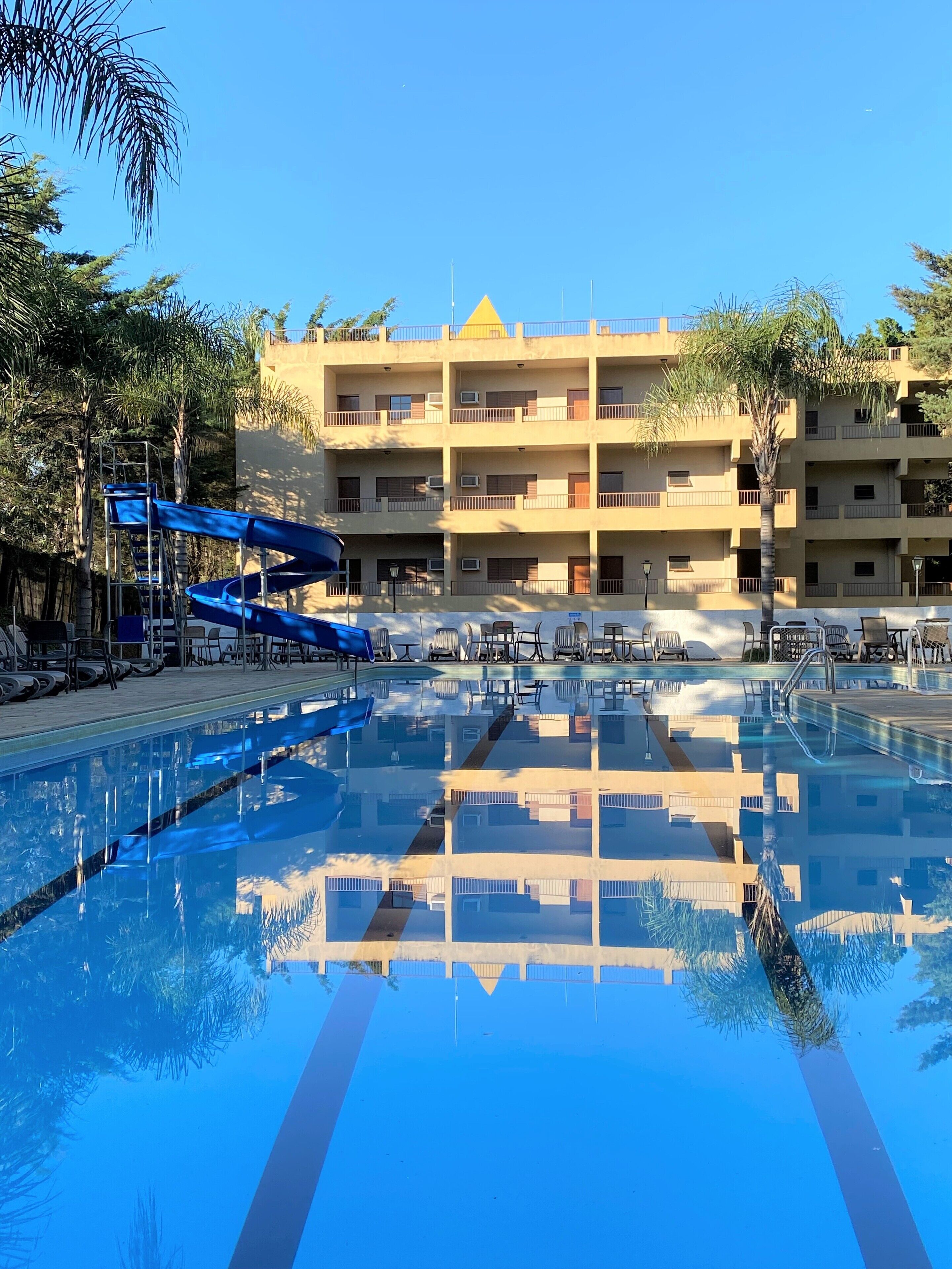 Una piscina con agua cristalina, rodeada de sillas de descanso y sombrillas. Al fondo, un edificio de varios pisos con balcones y unidades de aire acondicionado.