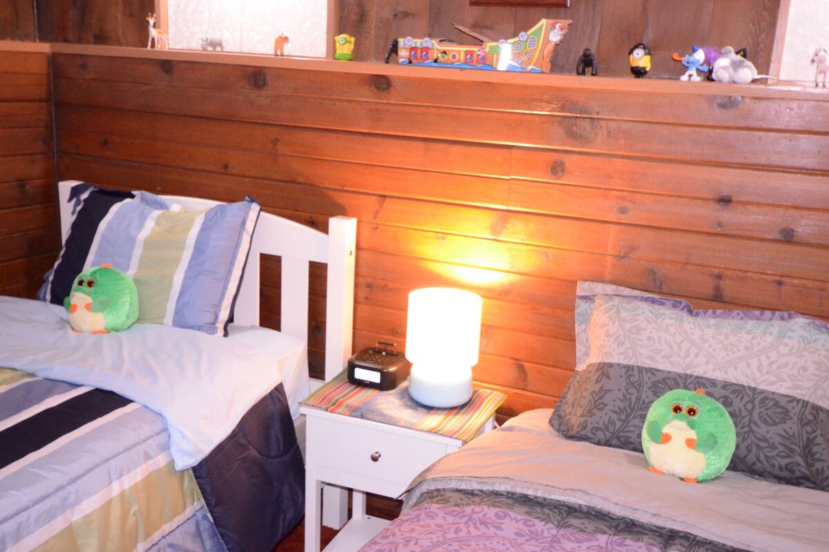 Een knus kamer in een blokhut met twee bedden, een nachtkastje met lamp en een plank met speelgoed.