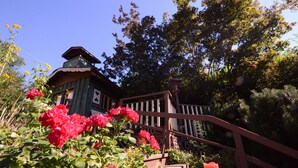 A rustic wooden building with a sloped roof, surrounded by lush greenery and vibrant red roses. - Fairy's Garden Chalet (West Kelowna)