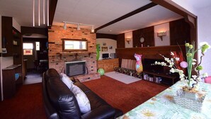 A living room with a brick fireplace, a leather sofa, a television, and a dining area with a table and chairs. - Fairy's Garden Chalet (West Kelowna)