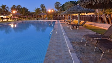 A swimming pool with lounge chairs and umbrellas, surrounded by palm trees and a wooden fence.