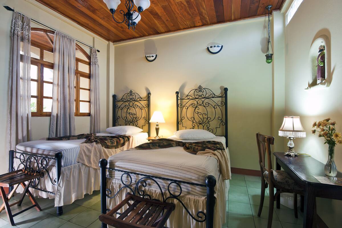 A room with two beds, a wooden ceiling, a table with a lamp, and a chair.
