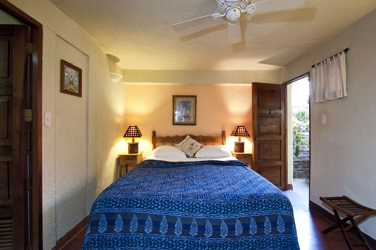 A bedroom with a large bed, two bedside lamps, a ceiling fan, and a door leading to an outdoor area.