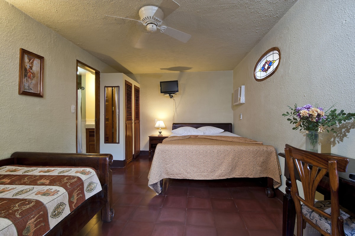 A hotel room with a bed, a desk, a chair, a ceiling fan, a TV, and a window with a stained glass design.