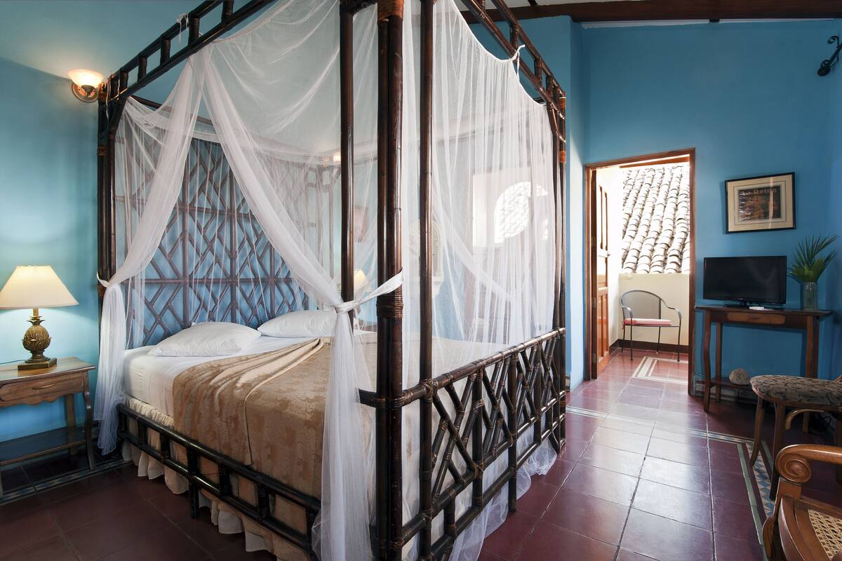 A four-poster bed with a canopy, a television on a stand, and a chair in a room with blue walls and a tiled floor.