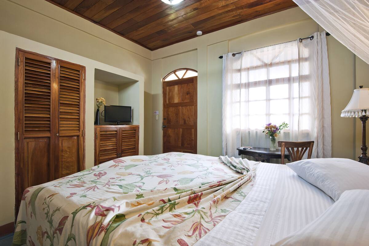 A bedroom with a floral bedspread, wooden wardrobe, a television, and a window with curtains.