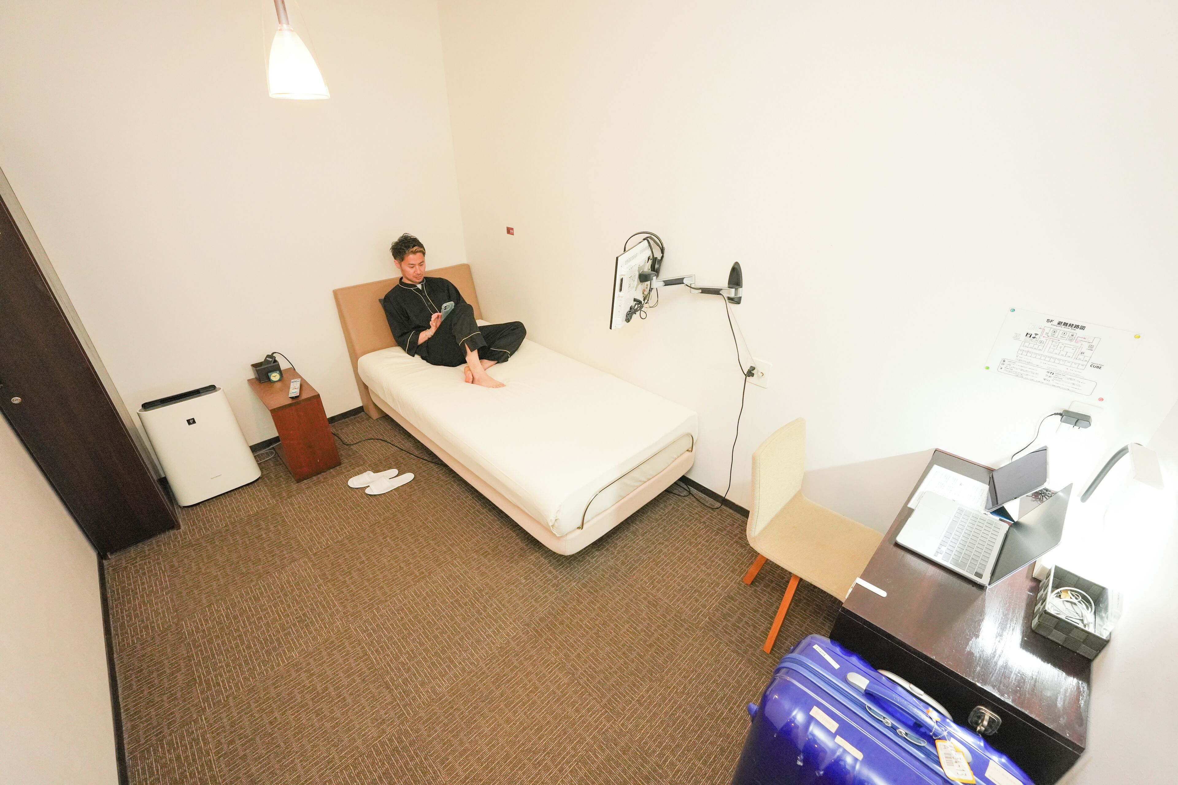 A hotel room with a bed, a desk with a laptop, a chair, a small desk lamp, a wall-mounted TV, and a suitcase.