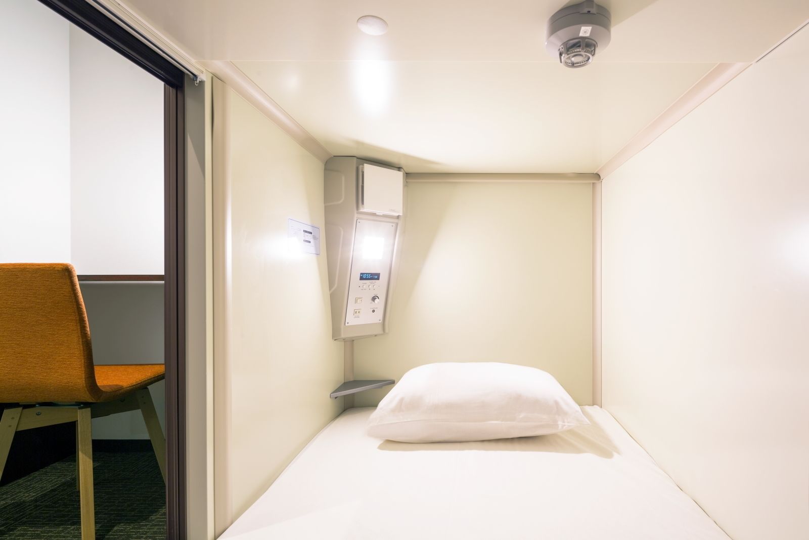 A small, compact room with a single bed, a chair, and a control panel.