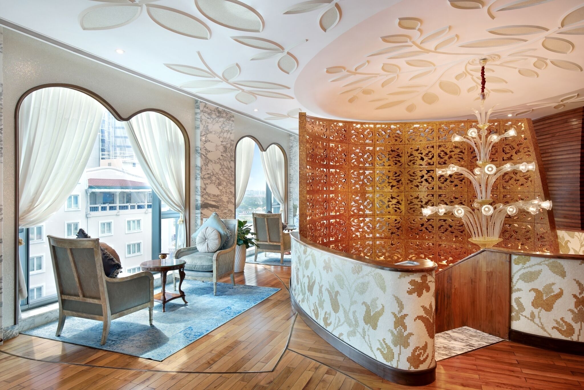 A modern hotel lobby with a curved reception desk, large windows with sheer curtains, and a decorative ceiling.