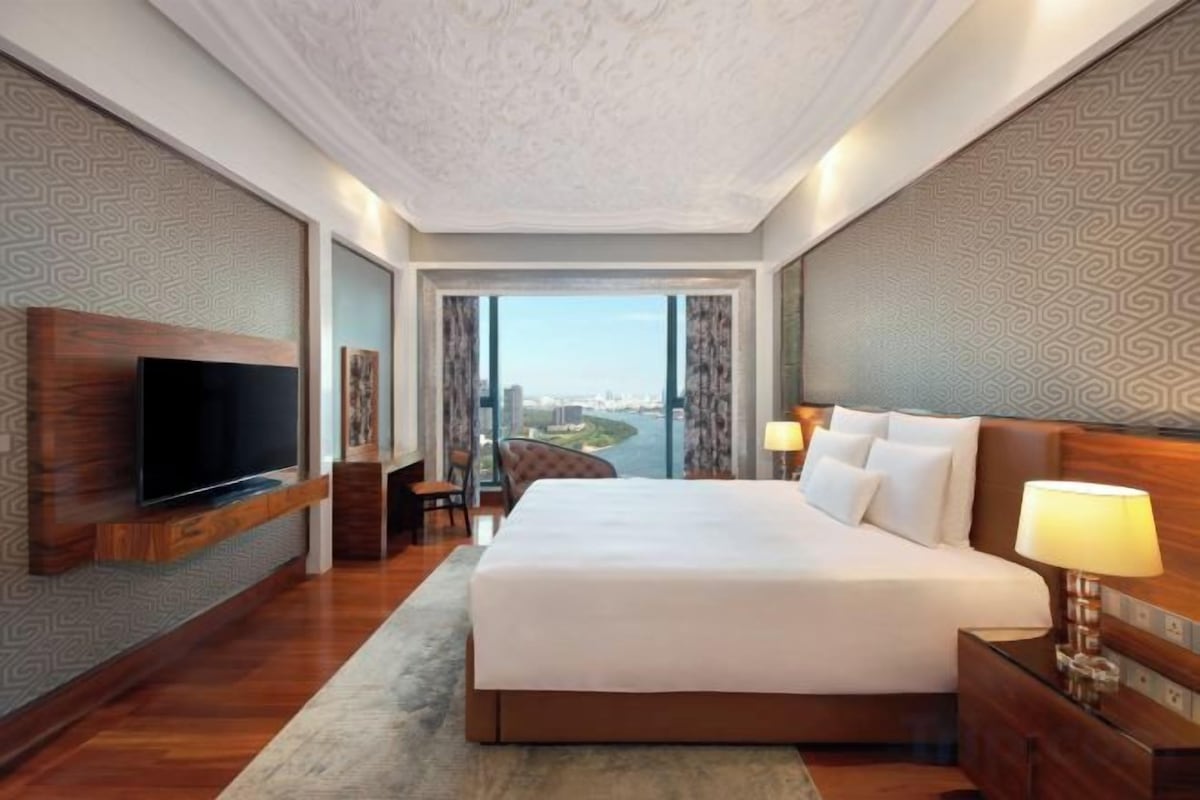 A hotel room with a large bed, a flat-screen TV, a desk, and a view of a cityscape through a large window.