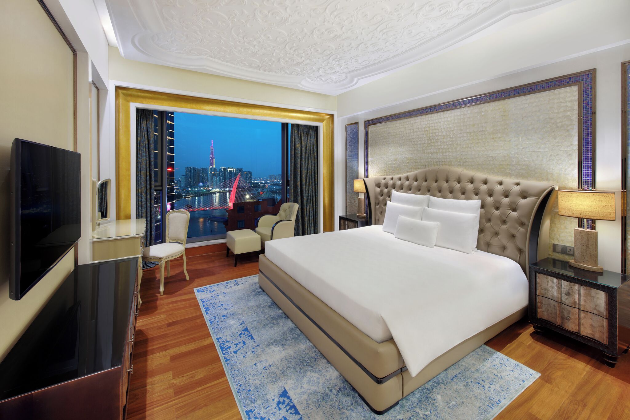 A spacious hotel room with a large bed, a television, a desk, and a view of a cityscape at dusk.