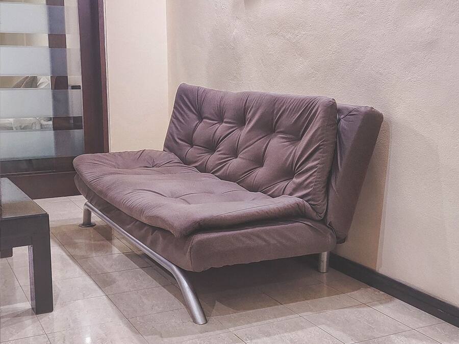 Un divano moderno con struttura metallica e seduta imbottita, appoggiato a una parete dalla superficie materica in una stanza con pavimento piastrellato.