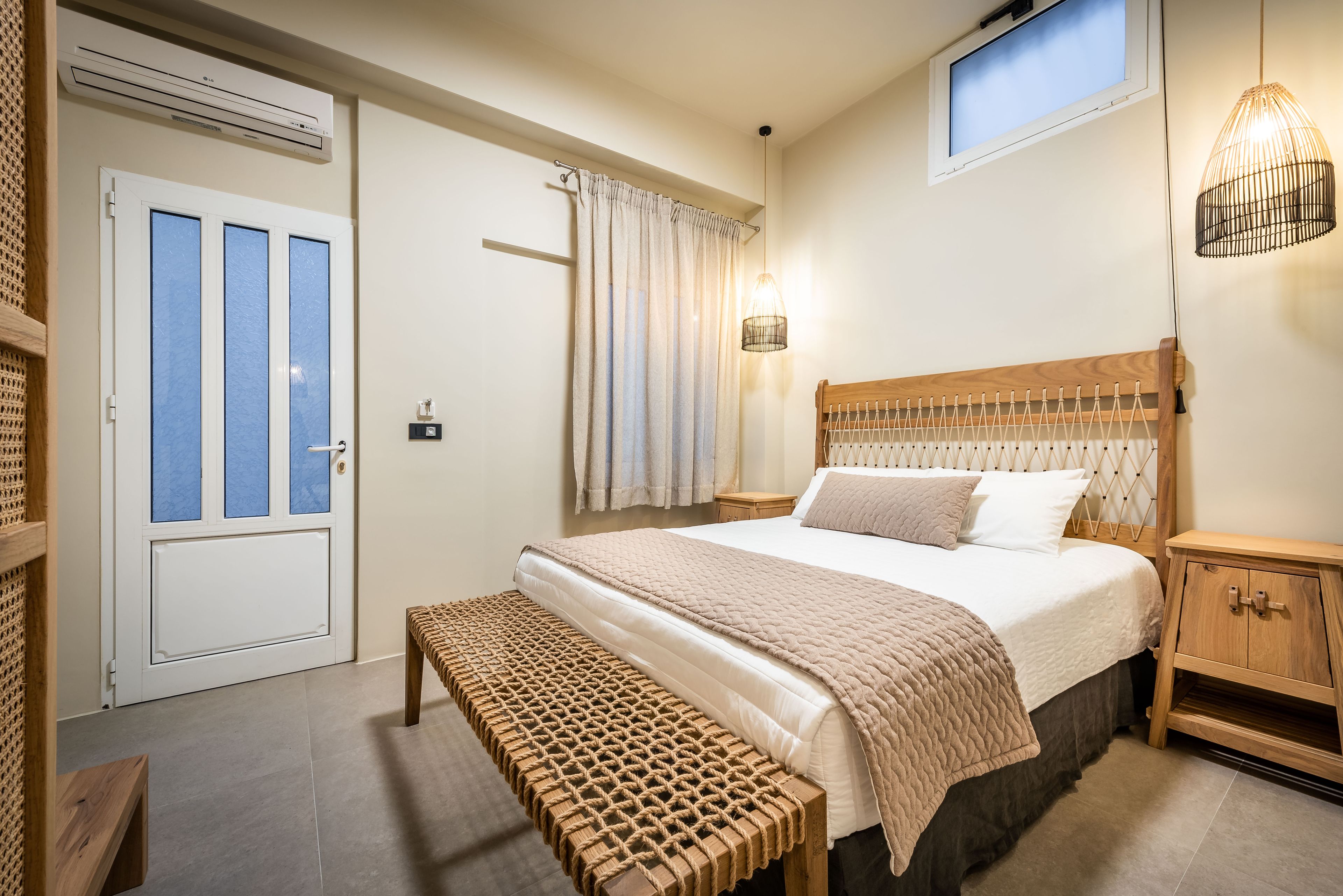 A bedroom with a bed, a bench, a wooden nightstand, a wall-mounted lamp, and a window with curtains.